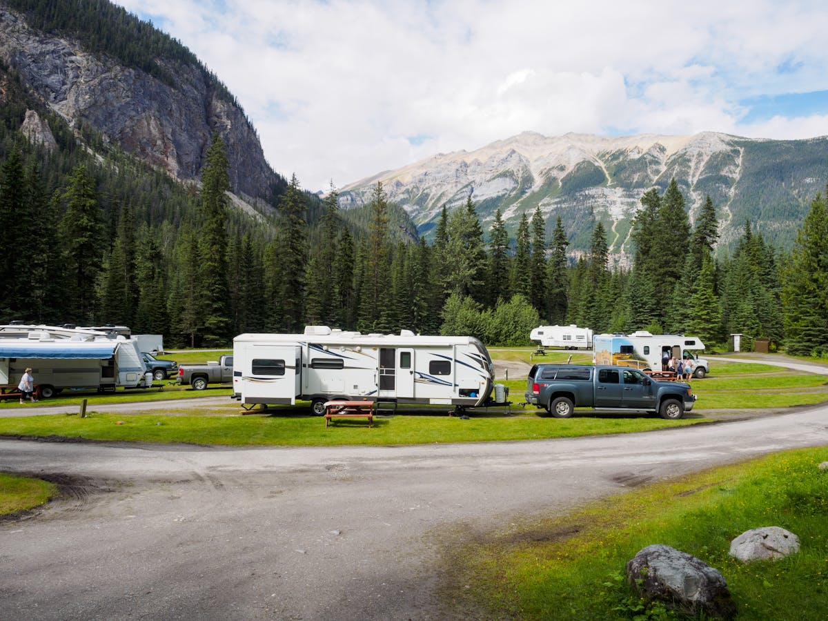 RVs and trailers at a mountain campsite, with forest and peaks behind.