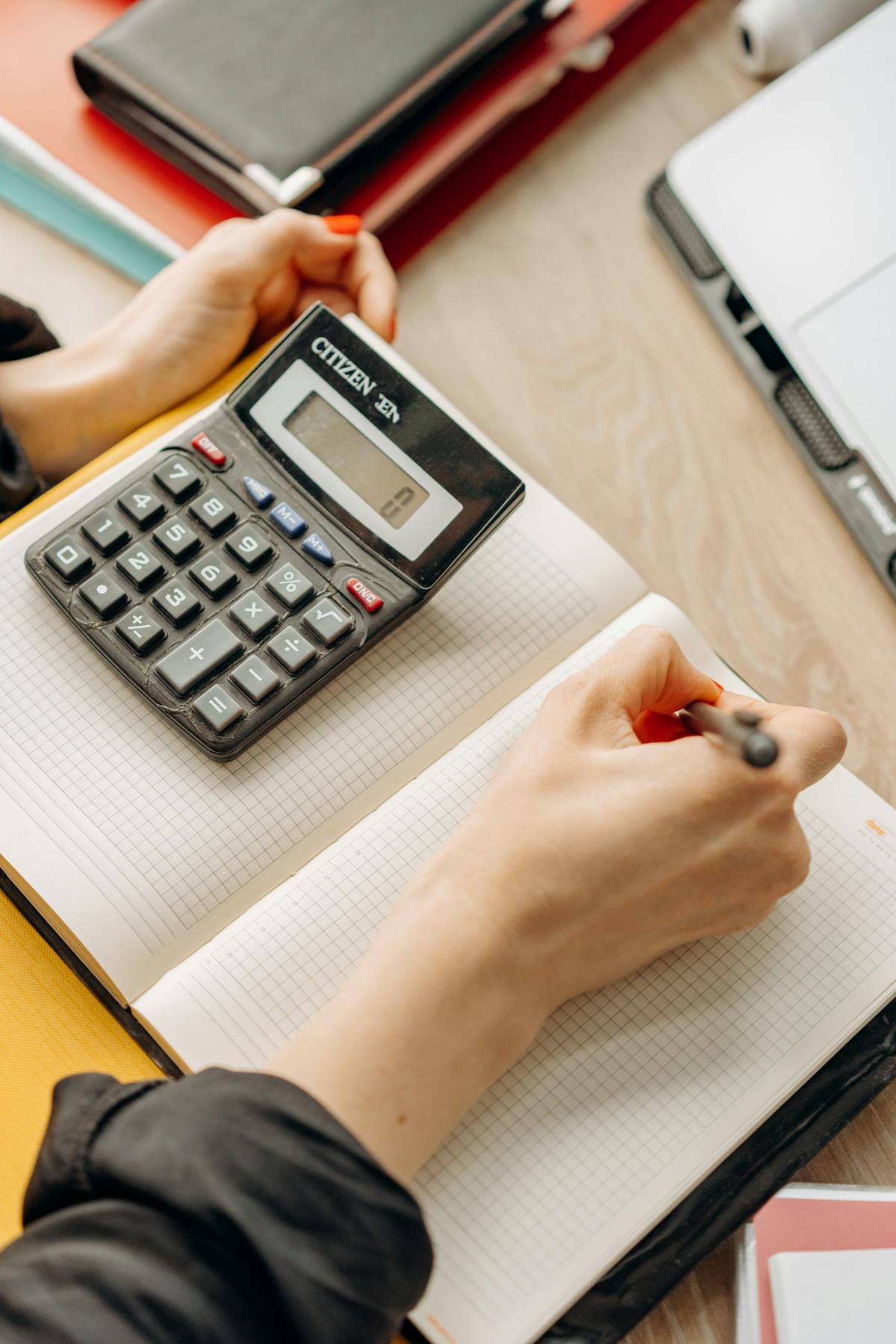 A hand writing in a notebook with a calculator on the desk.